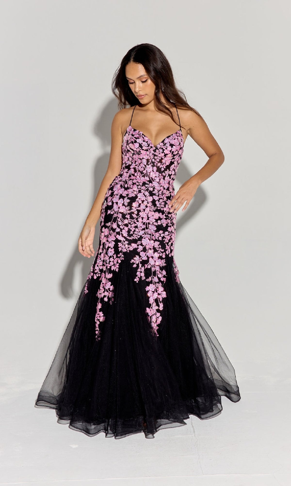 A woman stands against a plain light gray background wearing the Sequin-Floral Long Mermaid Prom Dress: Eureka 9696, a black floor-length gown with pink floral embroidery and a strappy back.